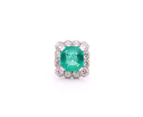 An emerald and diamond cluster ring; the rectangular cut-cornered emerald, in four claw mounts, within a border of twelve rou