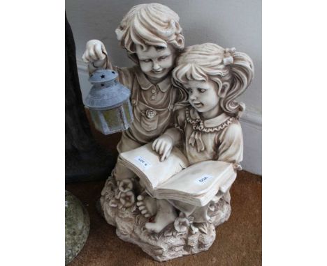 A CAST RESIN GARDEN FIGURE OF TWO CHILDREN READING BY LAMP LIGHT 