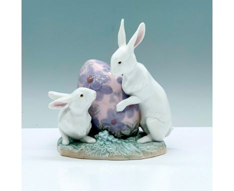 A figure of two white rabbits on a grass field inspecting a large pink egg that is decorated with purple flowers. Lladro back