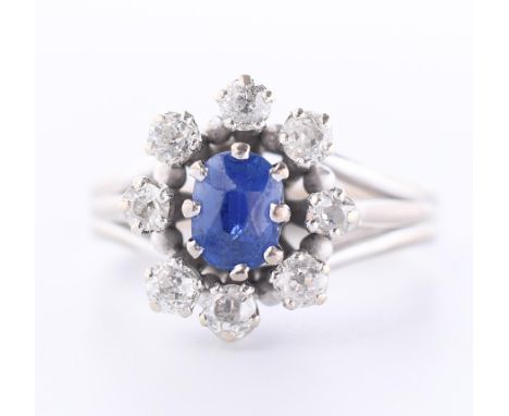 18 kt. Witgouden ring, centraal bezet met een ovale blauwe saffier (ca. 6 x 4,5 mm), in een entourage van briljant geslepen d