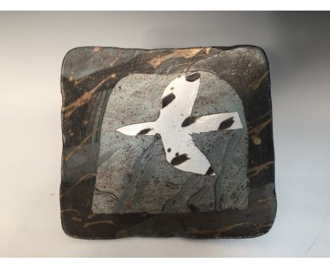 John Maltby (born 1936), a stoneware dish, the rounded square form raised on a square foot, decorated with a stylised bird in