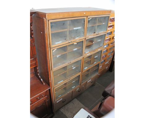 A light oak haberdashery cabinet, eight glazed lift up doors revealing four slide trays in each section, above three drawer b