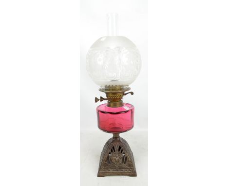 A cast iron oil lamp with etched globular shade, cranberry tinted reservoir and pierced floral decorated base, height includi