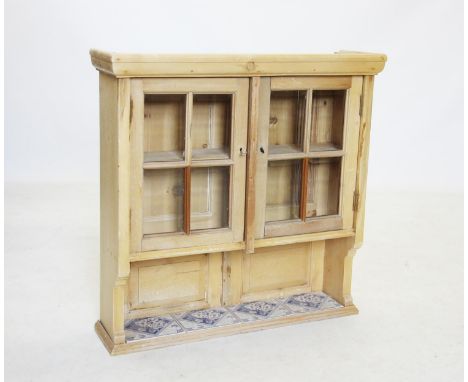 A glazed pine wall cupboard, early 20th century, the glazed doors above a single shelf inset with four blue and white tiles, 