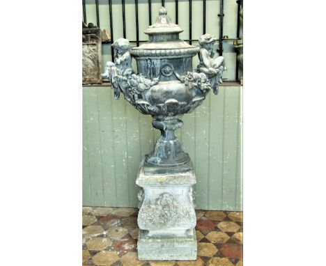 A weathered lead trophy shaped garden urn with removable lid, fruiting swags, seated cross legged cherubs, acanthus and furth