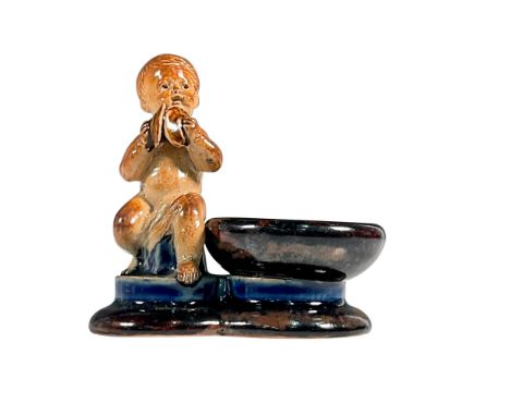 A rare stoneware open salt cellar modeled as a young boy playing cymbals seated beside a circular dish, raised on a shaped ba