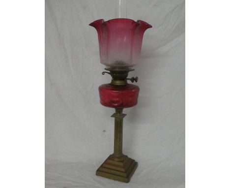 An Edwardian brass column oil lamp with cranberry-tinted glass reservoir and cranberry-tinted etched glass shade (shade damag