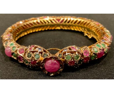 A gold plated hinged pierced openwork bangle encrusted overall with low grade cabochon rubies and facet cut emeralds, etc. 