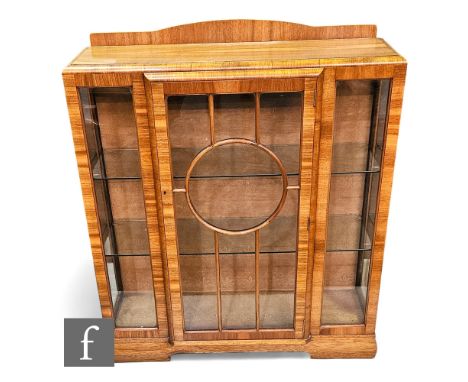 A 1930s Art Deco walnut breakfront display cabinet enclosed by a circular bar glazed door below a shaped shelf on plinth base