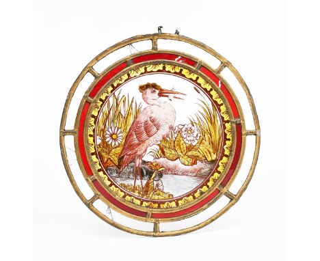 An Aesthetic Movement stained glass roundel, painted with an anthropomorphic crane bird standing on a river bank with a fish 