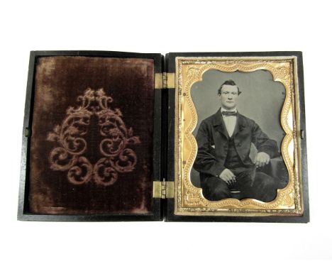 A Victorian tintype photograph of a seated gentleman wearing a frock coat, hand-tinted, under gilt preserver in a figural gut