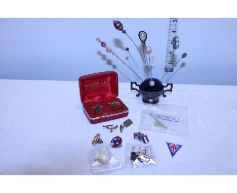 A white metal pin cushion with a selection of vintage hat pins, gold plated cufflinks and badges 