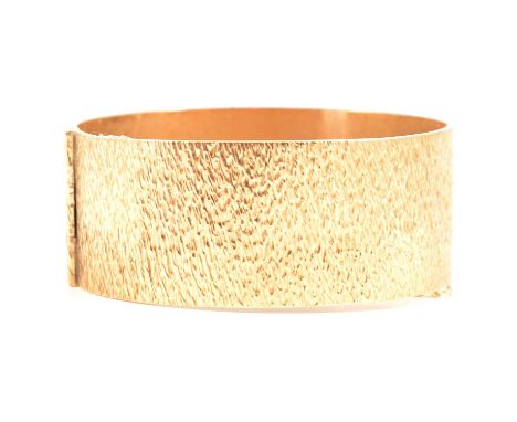 A 9 carat yellow gold bark texture half-hinged bangle, 15mm wide, Birmingham 1970, with safety chain, internal dimensions 58 
