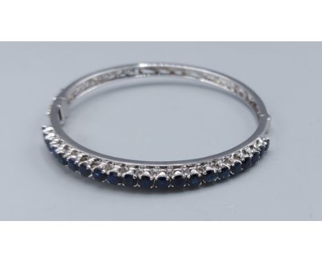 A white metal bangle set with a row of twenty one sapphire, claw set, 12.9gms, 6.5cms diameter 