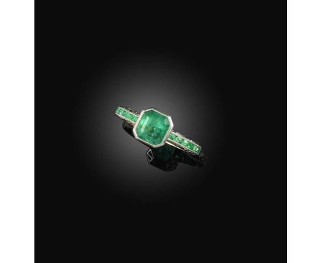 An emerald ring, millegrain-set with a step-cut emerald, to a platinum band engraved with foliate decoration and set with a l