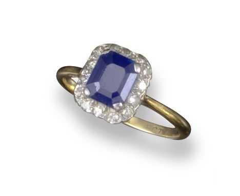 A sapphire and diamond cluster ring, the emerald-cut sapphire is set within a surround of single-cut diamonds in platinum and