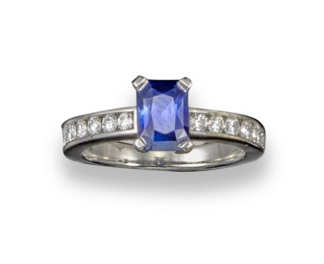 A sapphire and diamond ring, the emerald-cut sapphire is set within channel-set diamond shoulders in white gold, size M