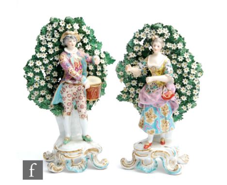 A pair of early 20th Century continental figures of a lady and gentleman stood before a floral bocage, he playing a drum, she