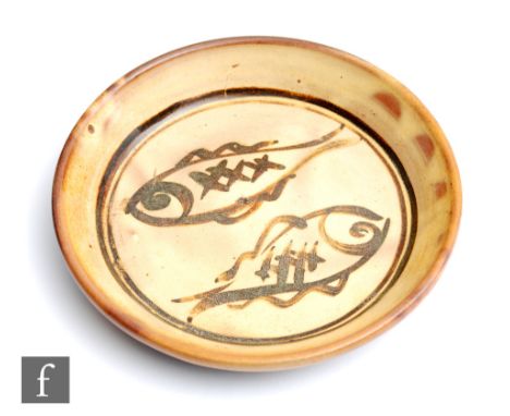 A small Winchcombe Pottery studio pottery dish decorated by Patrick Groom with two brown painted fish against the ochre groun