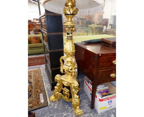 A CARVED GILT AND EBONISED STANDARD LAMP WORKED WITH MASKS AND FOLIAGE ABOVE THREE PAW FEET, THE SHADE WITH FLORAL APPLIQUES