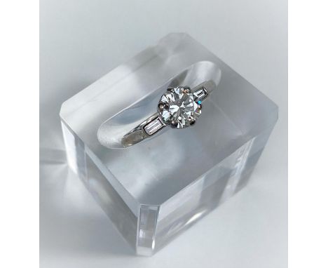 A diamond set ring with central bright cut stone and small baguette cut stone to either shoulder, on white metal shank (6.4mm