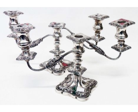 A pair of silver twin branch candelabra with detachable nozzles, ornate cast and embossed decoration, set on loaded shaped ba