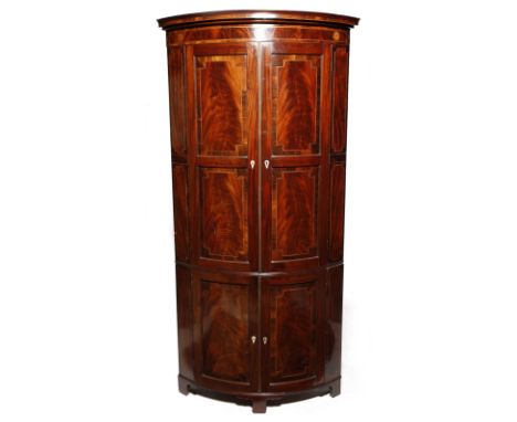 λ  A late George III mahogany bowfront standing corner cupboard, in two sections, inlaid with stringing and with rosewood ban