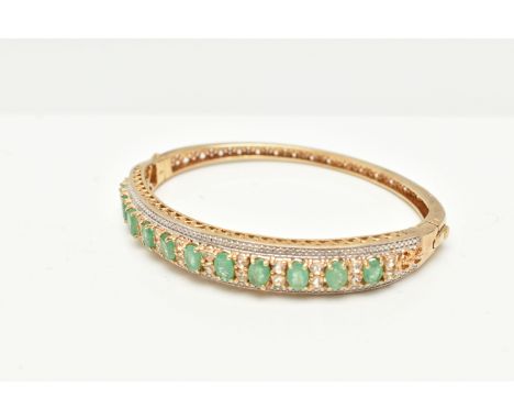 A GILT HINGED BANGLE, designed with a row of eleven oval cut emeralds, each in a four claw setting, interspaced with colourle