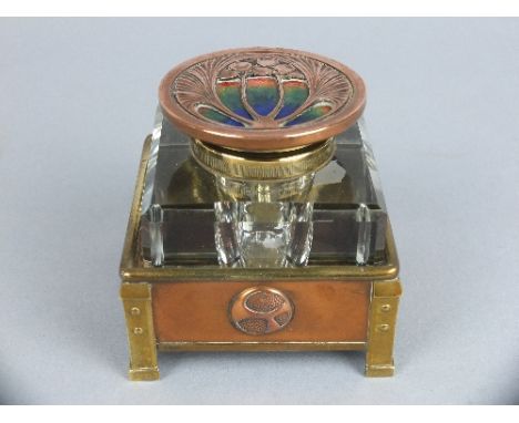 AN ARTS & CRAFTS STYLE SQUARE GLASS INKWELL, hinged circular copper cover with cast foliate motif and enamel inset, loosely f