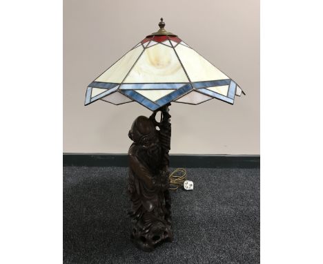 A carved wooden lamp base modelled as a Chinese elder with staff with a leaded glass shade 