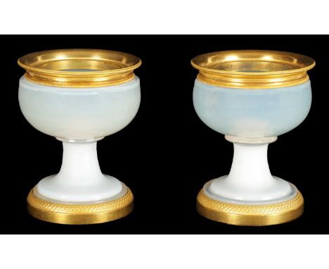 A PAIR OF EARLY 19TH CENTURY FRENCH ORMOLU MOUNTED WHITE OPALINE GLASS CUPS of squat bulbous form raised on pedestal bases wi