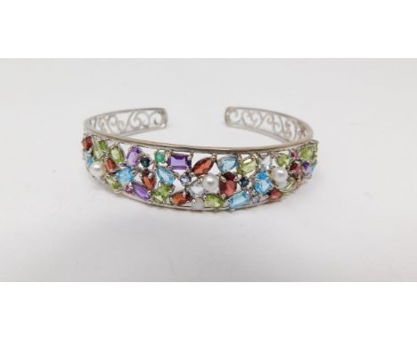 A multi gemstone and silver scrolling design pierced bangle. Set with forty eight mixed cut gemstones in open back claw setti