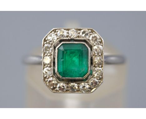 A white metal cluster ring. Principally set with a rectangular cut emerald estimated to weigh 0.65cts. Surrounded by grain se