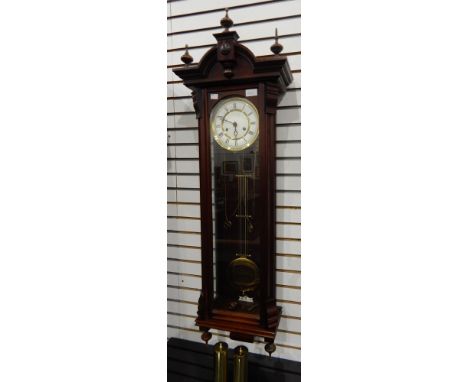 A Vienna regulator-style reproduction stained wood wall clock having turned finials to the arched pediment, large brass compe