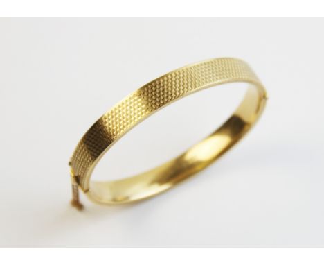 A 9ct gold hinged bangle, of oval form with engine turned decoration to exterior, with tongue and box snap clasp and safety c