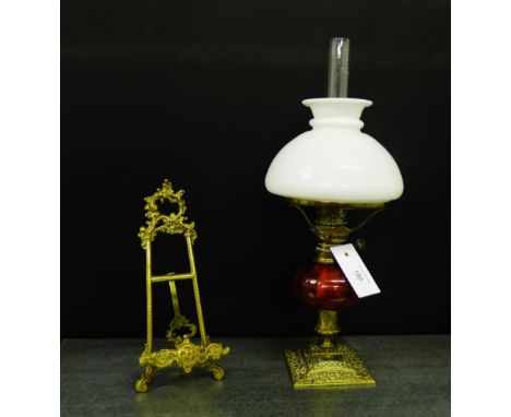 A brass table oil lamp with a cranberry glass well and a white opaque glass shade, together with a cast brass easel stand, ta