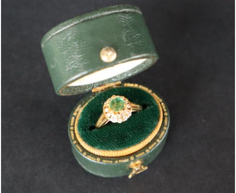 An emerald and diamond ring set with a central circular faceted emerald surrounded by two tiers of round brilliant cut diamon