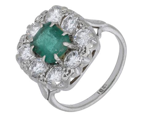 An emerald and diamond cluster ring, set with a step-cut emerald within a surround of brilliant-cut diamonds, between engrave