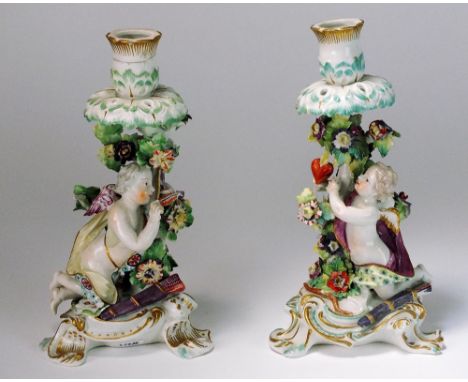 A pair of Derby candlesticks depicting Cupid, one extracting an arrow and the other pointing towards a flaming heart, on scro