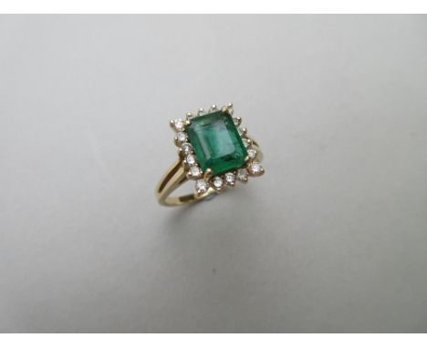 An emerald and diamond ring, the emerald cut emerald four claw set in a border of eighteen round brilliant cut diamonds, to a