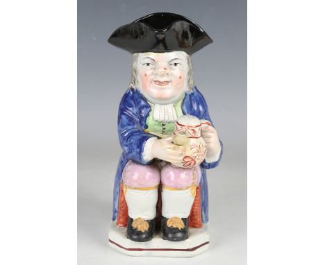 A pearlware Toby jug, circa 1820, modelled seated with a warty face, holding a jug of ale and a pipe, wearing a blue frock co