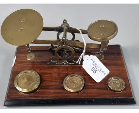 Set of brass postal scales with one, two and four ounce brass weights on a stained wooden base.  20cm wide approx.  (B.P. 21%