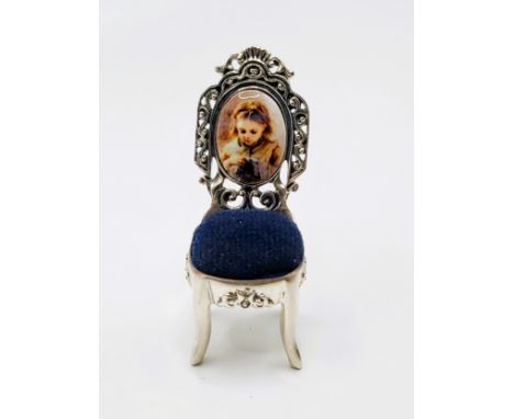 A silver pin cushion in the form of a chair with a portrait of a young girl sewing on the back. Stamped sterling silver. 4.5c