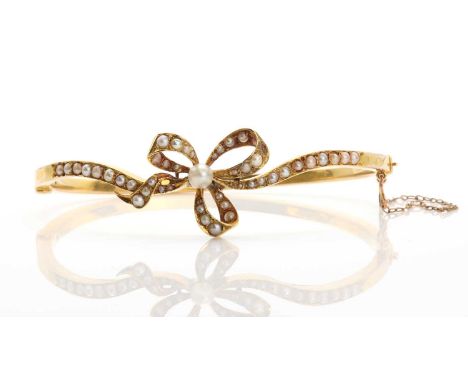 An Edwardian split pearl set hinged bangle, c.1905,  with an open bow centrepiece, peg set with 4.10mm pearl, to lines of gra