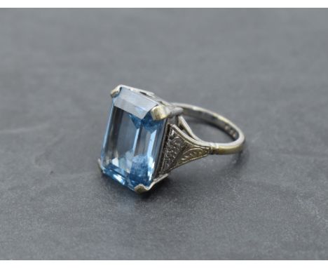 An oversized dress ring having a rectangular cut blue topaz in a four claw basket mount to embossed shoulders on a white meta