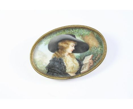 A PORTRAIT MINIATURE BROOCH depicting a young girl in a large hat reading a book under a tree, in gilt metal frame, 6 x 4cm