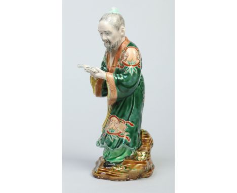 An early 20th century Chinese famille vert figure of a scholar. Wearing a long flowing robe painted with fish, reading from a