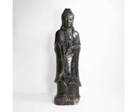 A large Chinese spinach jade carving, of Guanyin, standing with her eyes downcast in a serene expression, holding a small vas