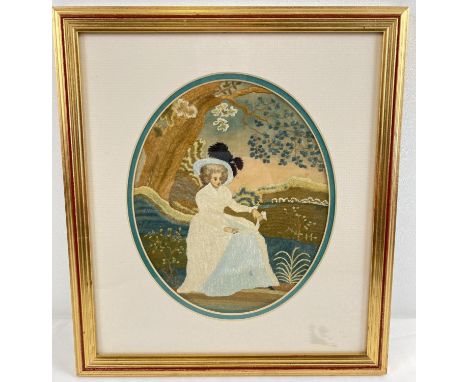 A framed and glazed fine silk embroidered panel of a Georgian lady sitting beneath a tree. In brown, cream and teal colourway