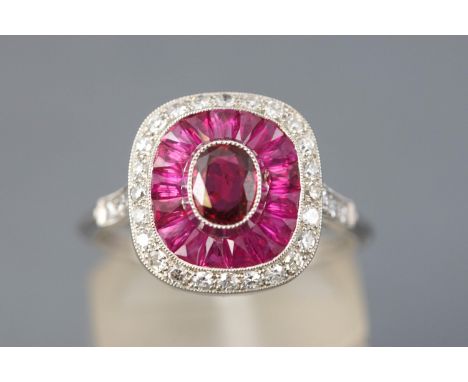 A white metal cluster ring. Principally set with an oval faceted cut ruby measuring approximately 5mm x 4mm. Surrounded by ca